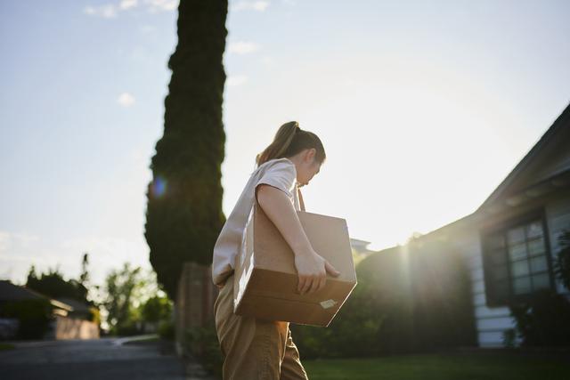 Person in tan outfit carrying a sealed cardboard box past a tall cypress tree at sunset