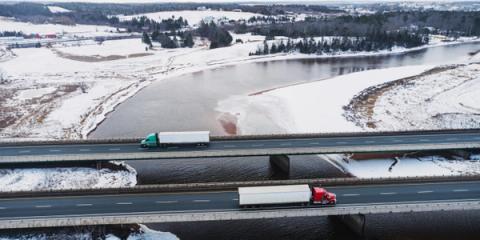 Aerial view of two semi-trucks crossing parallel highway bridges over an icy river in a snowy landscape