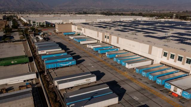 Aerial view of a large warehouse with rows of blue Amazon-branded trailers parked at loading docks and solar panels on the roof