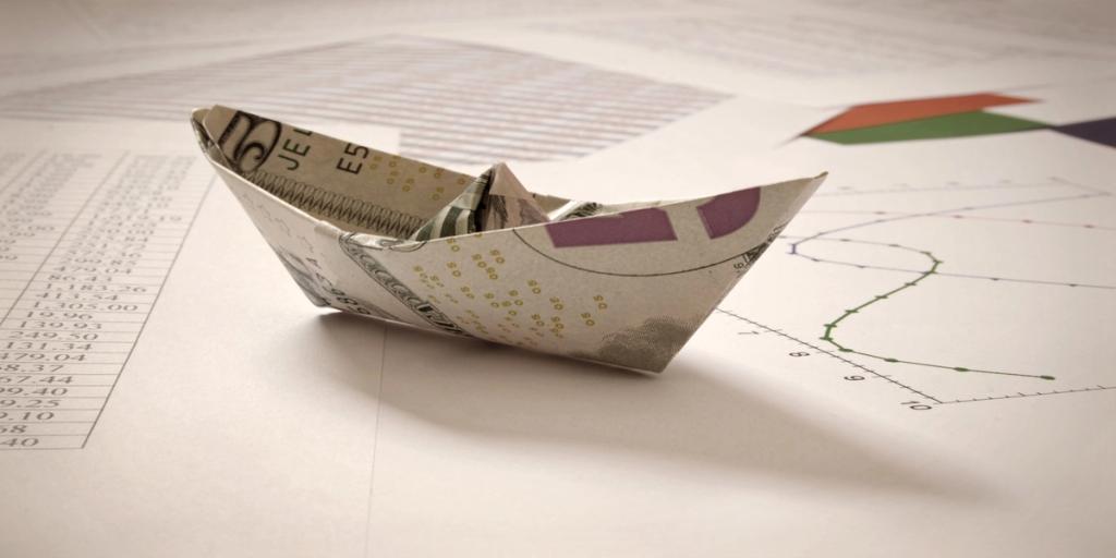 Paper boat folded from a banknote sitting on printed financial spreadsheets and charts