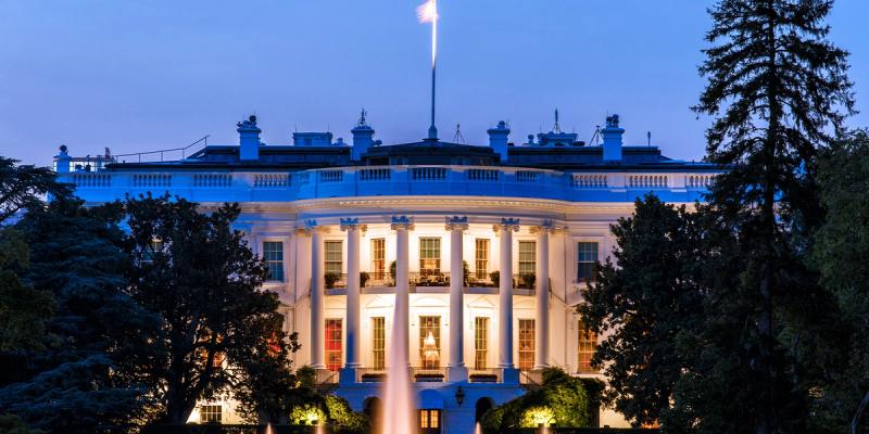 The South Portico of the White House lit up at dusk with a fountain and trees in front