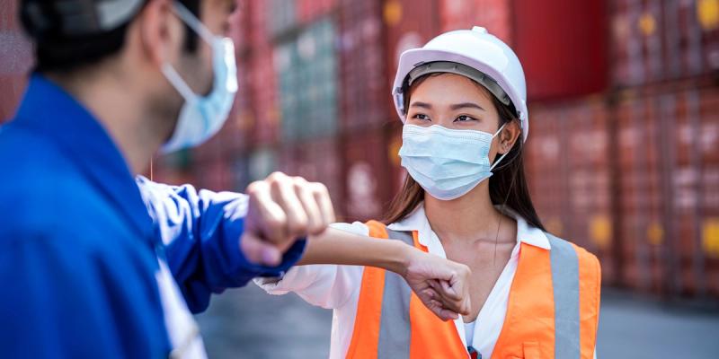 Two masked workers in a container yard bump elbows; woman wears a hard hat and orange safety vest.