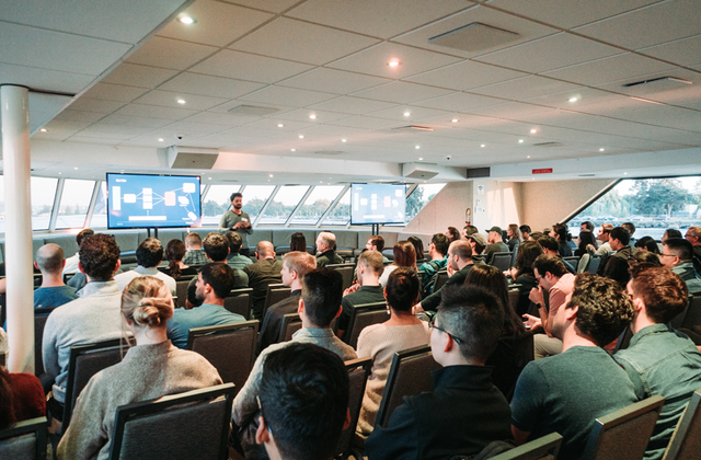 Audience seated in rows facing a presentation screen inside a windowed event space aboard a vessel