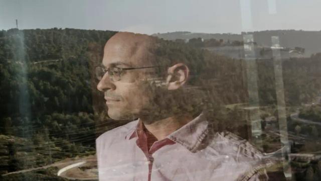 Profile portrait of a bald man in glasses and a pink shirt, seen through a glass window reflecting a forested hillside