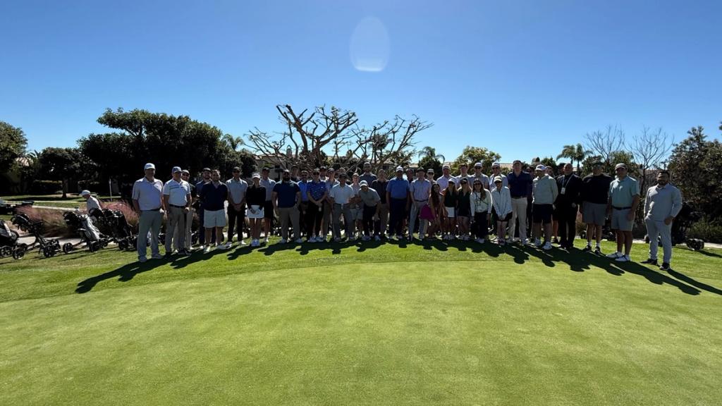 Large group of golf-event attendees posing on a sunlit putting green under a clear blue sky