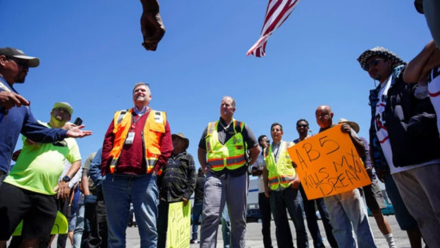 Group of people gathered outdoors at a rally, men in safety vests beside an American flag, one holding a handmade cardboard sign