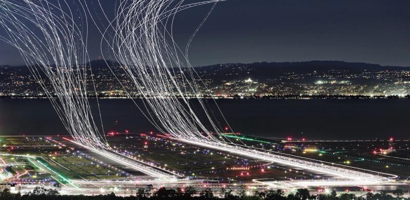 Long-exposure night photo of light trails from aircraft taking off over an airport, with a city skyline across the bay