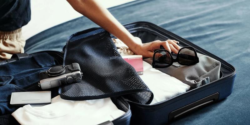 Open suitcase on a bed with a camera, passport, sunglasses and clothes; hand reaching in to pack