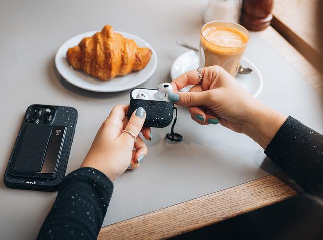 Hands opening a speckled Mous earbud case at a cafe table with a croissant, latte and phone