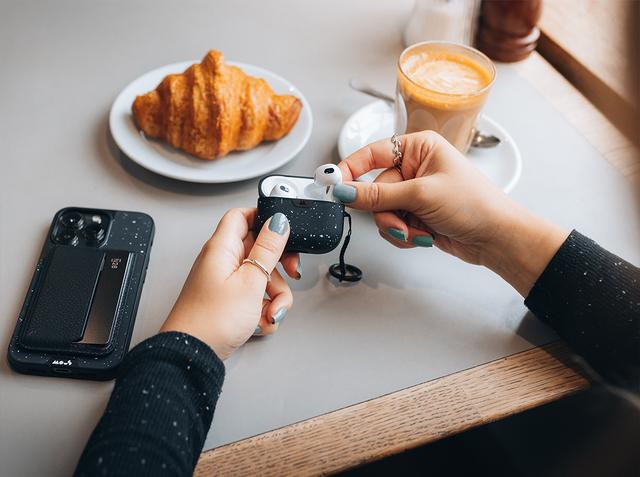 Hands opening a speckled Mous earbud case at a cafe table with a croissant, latte and phone