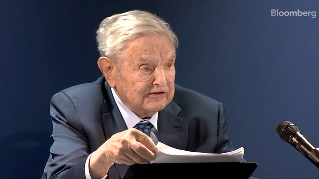 Bloomberg broadcast still of an elderly man in a dark suit speaking at a panel, holding papers, with Bloomberg watermark