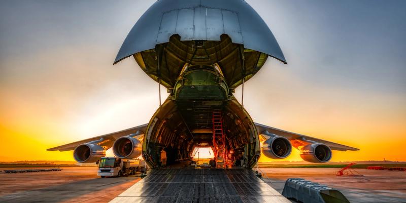Wide-body cargo plane with raised nose cone loading freight on an airport apron at sunset