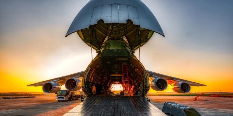 Wide-body cargo plane with raised nose cone loading freight on an airport apron at sunset