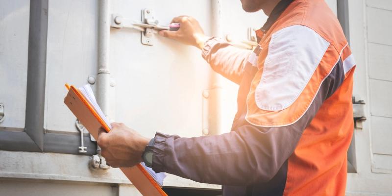 Worker in an orange high-visibility jacket holding a clipboard while opening a shipping container door
