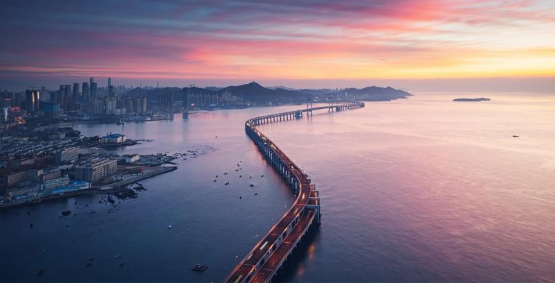 Aerial view of a long curving sea bridge linking a coastal city to the open water at sunset