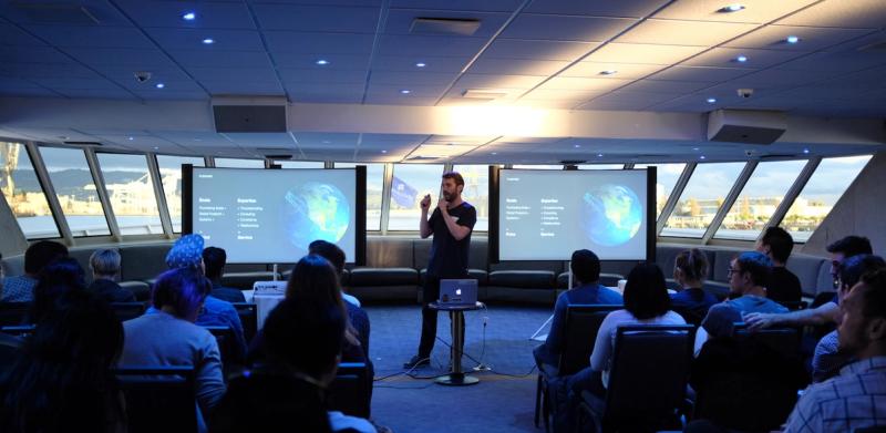Man presents to a seated audience aboard a vessel, with two projection screens showing Flexport slides and a port view through windows.