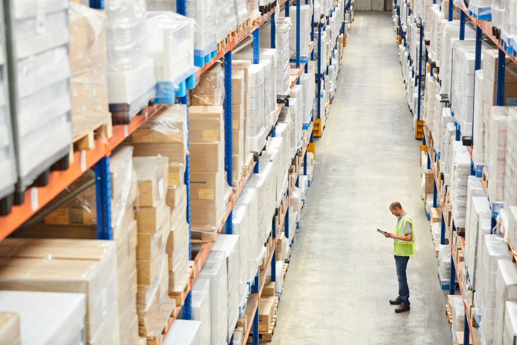 Warehouse worker checks inventory on tablet among tall shelving racks.