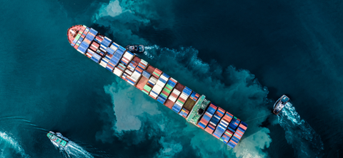 Overhead aerial photo of a laden container ship under escort by tugboats across dark teal water.