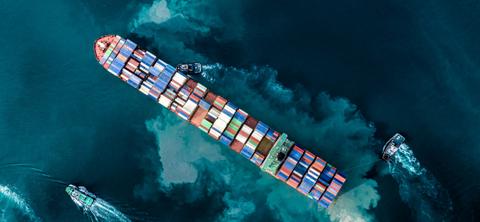 Overhead aerial photo of a laden container ship under escort by tugboats across dark teal water.
