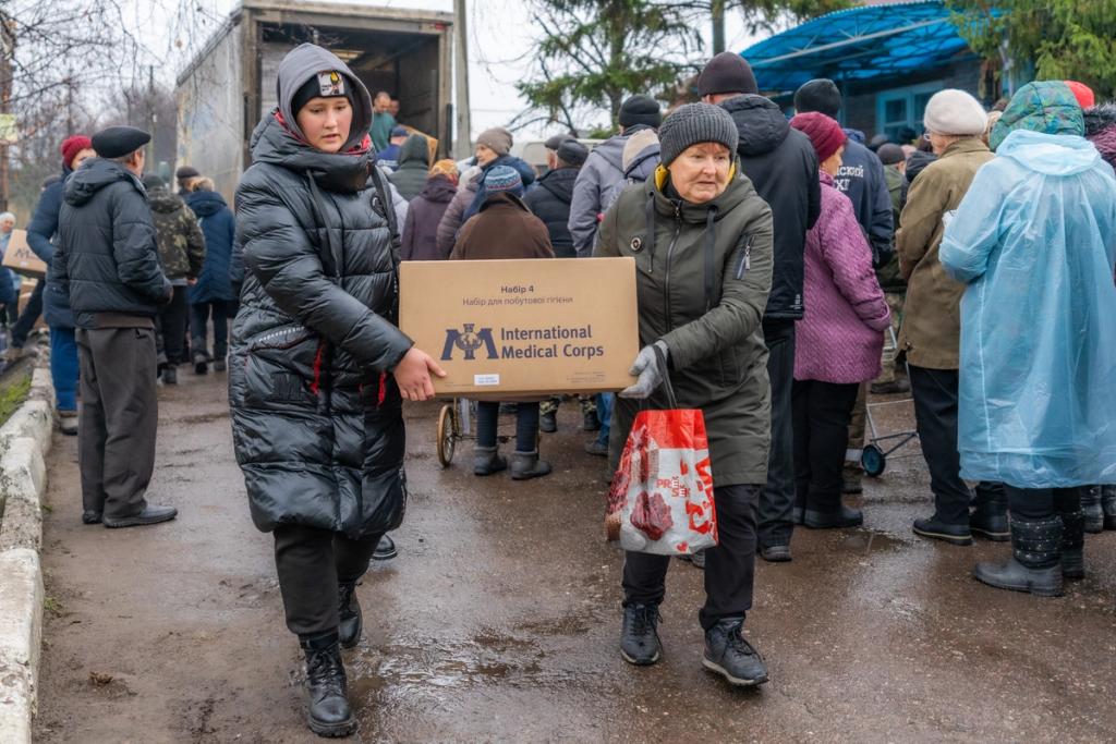 Two women carry an International Medical Corps aid box past a crowd queuing at a relief distribution truck