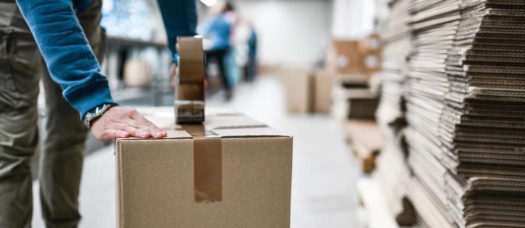 Warehouse worker sealing a cardboard shipping box beside stacks of flattened cartons