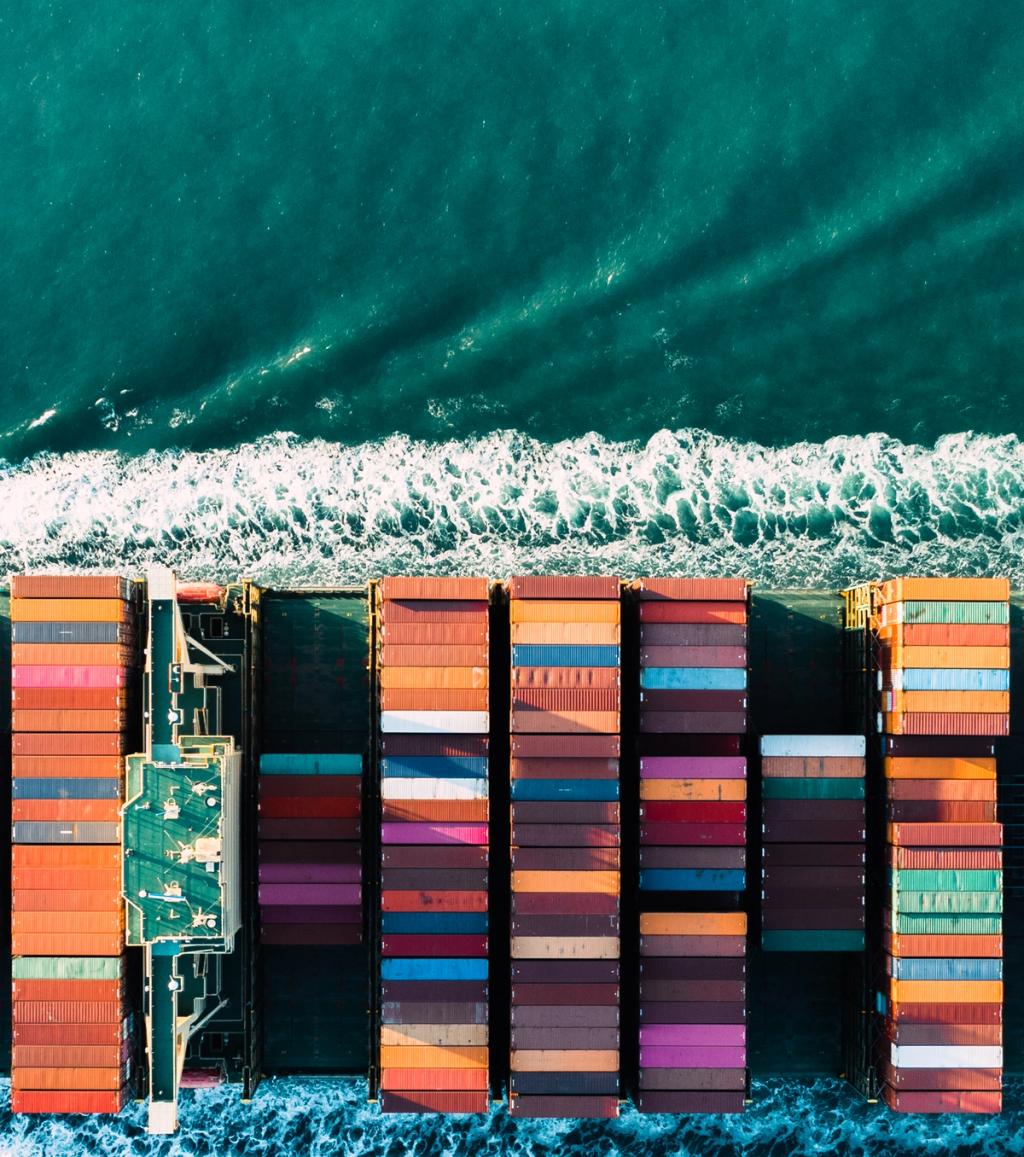 Overhead aerial view of a fully loaded container ship cutting through deep blue ocean water leaving a white wake