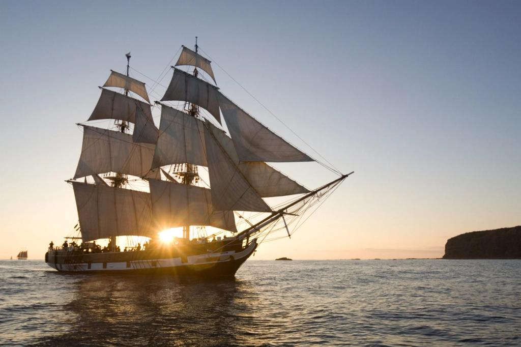 Square-rigged tall sailing ship under full sail on calm water with the sun setting behind its hull