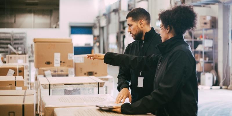 Two warehouse workers in black jackets examining a tablet and pointing among stacked pallets and boxes
