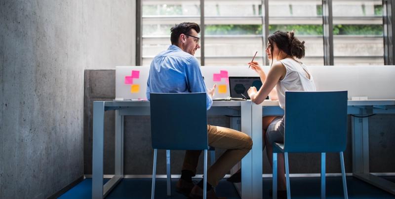 A man and woman in business attire talk across a desk with sticky notes and a laptop in a modern office