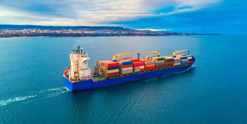 Blue container ship with crane sailing on open water near a coastline at dusk
