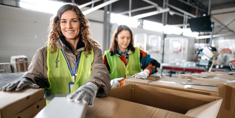 Two women in high-visibility vests and gloves packing products into cardboard boxes in a warehouse