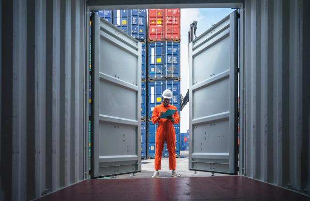 Worker in orange coveralls and hard hat writing on a clipboard, framed by open container doors with stacked containers behind