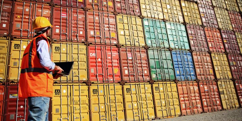 Worker in hard hat and orange vest holds a laptop facing a tall wall of stacked containers
