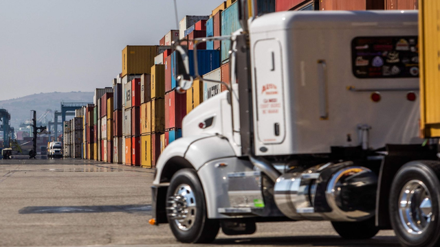 White semi-truck cab parked at a port beside rows of stacked shipping containers