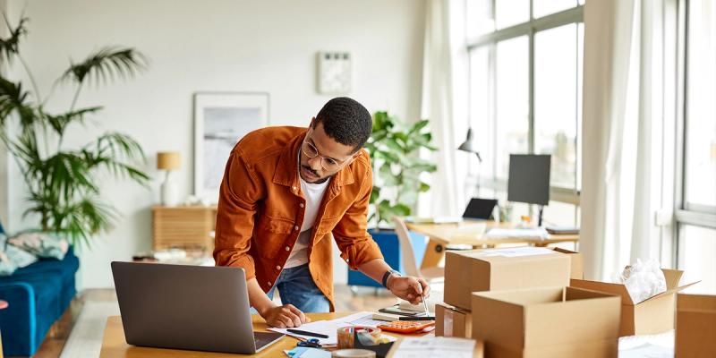 Man in an orange jacket and glasses working at a laptop with shipping boxes on a home office table