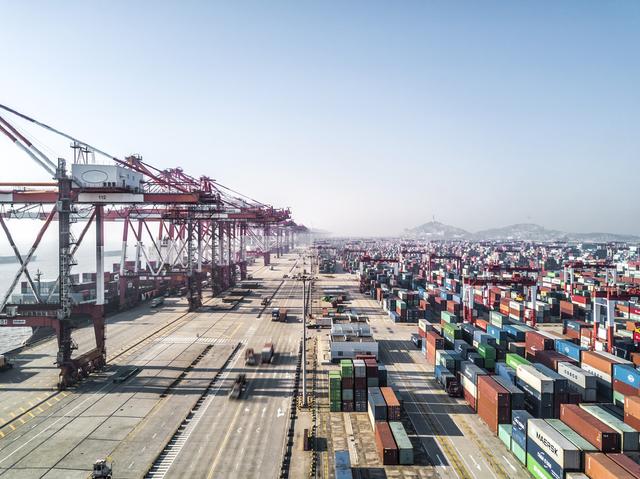 Aerial view of a container port with red gantry cranes and rows of stacked containers stretching to hills.