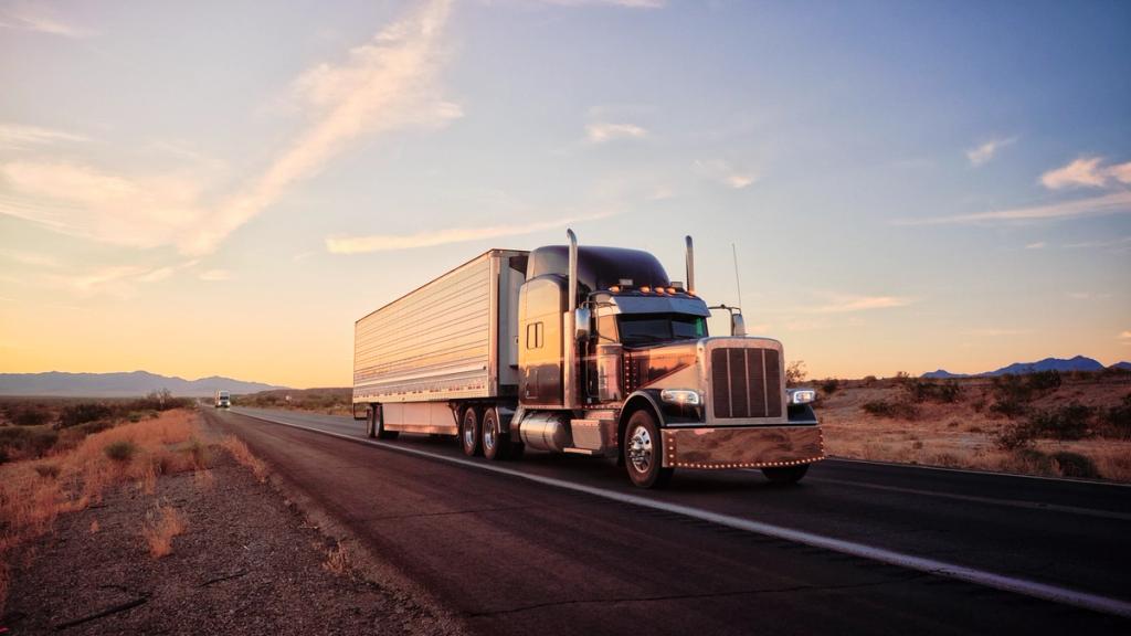 Semi-truck with silver trailer driving down a desert highway at sunset with mountains in the distance