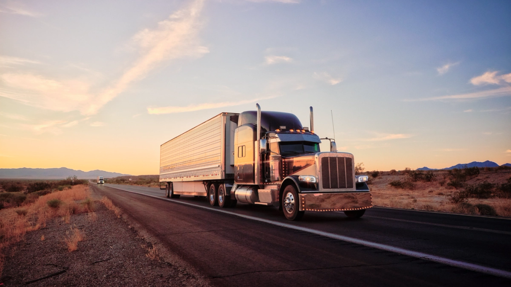 Semi-truck with silver trailer driving down a desert highway at sunset with mountains in the distance