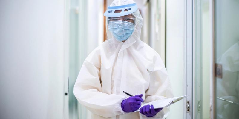 Person in white protective suit, face shield, mask and purple gloves holding a clipboard in a corridor