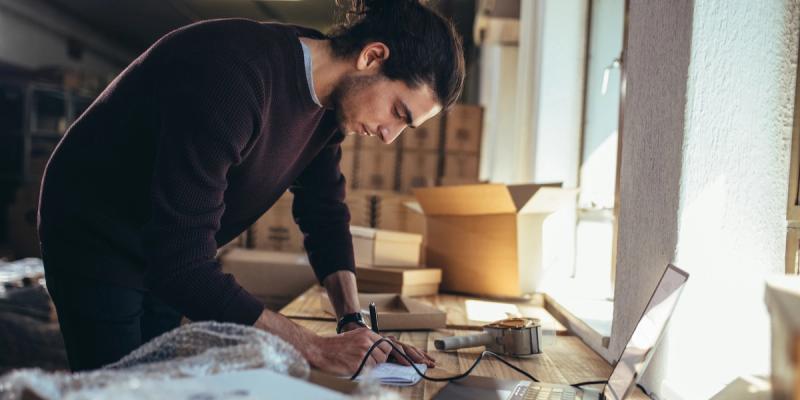 Man with a bun bends over a workbench writing in a notebook amid cardboard boxes and packing tape.