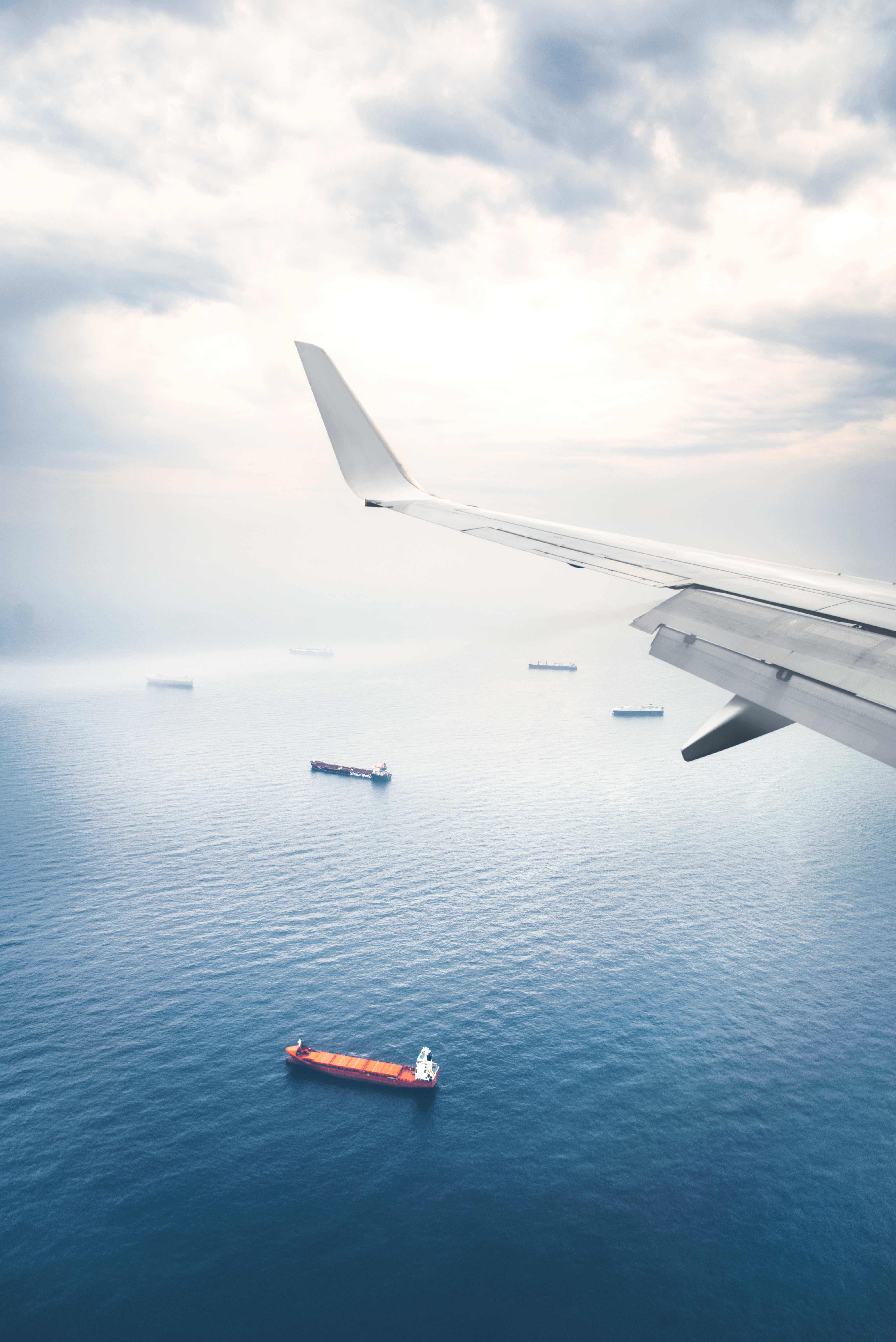 Aircraft wing extends over open ocean with cargo ships visible below.