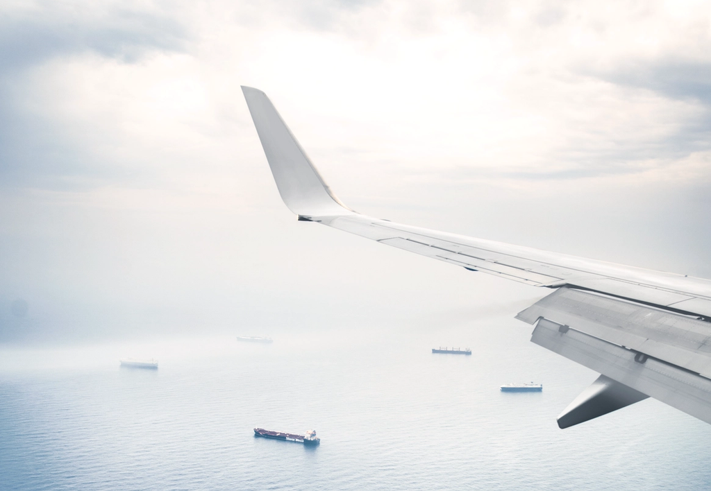 Aircraft wing extends over open ocean with cargo ships visible below.