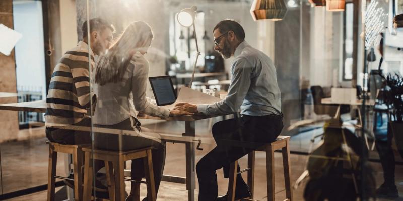 Three colleagues collaborate over a laptop at a high table in a modern office, seen through a glass partition.