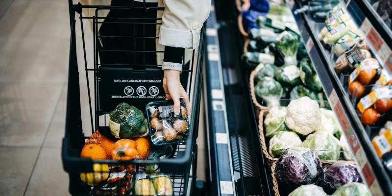 Shopper's hand placing packaged produce into a cart in a supermarket's vegetable aisle