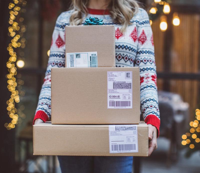 Person in a holiday sweater holding a stack of labeled parcels in front of string lights