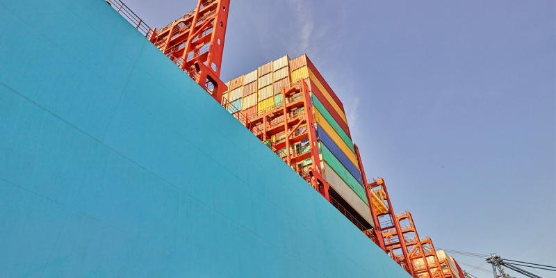 Low-angle view of a teal container ship hull with colorful stacked containers and orange crane structures above