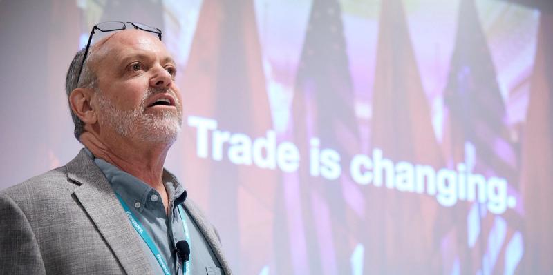 Bearded man speaks before a screen reading 'Trade is changing.' with U.S. flags projected
