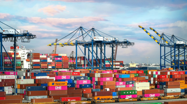 Container port at dusk with stacked shipping containers and rows of cranes under a pink and blue sky