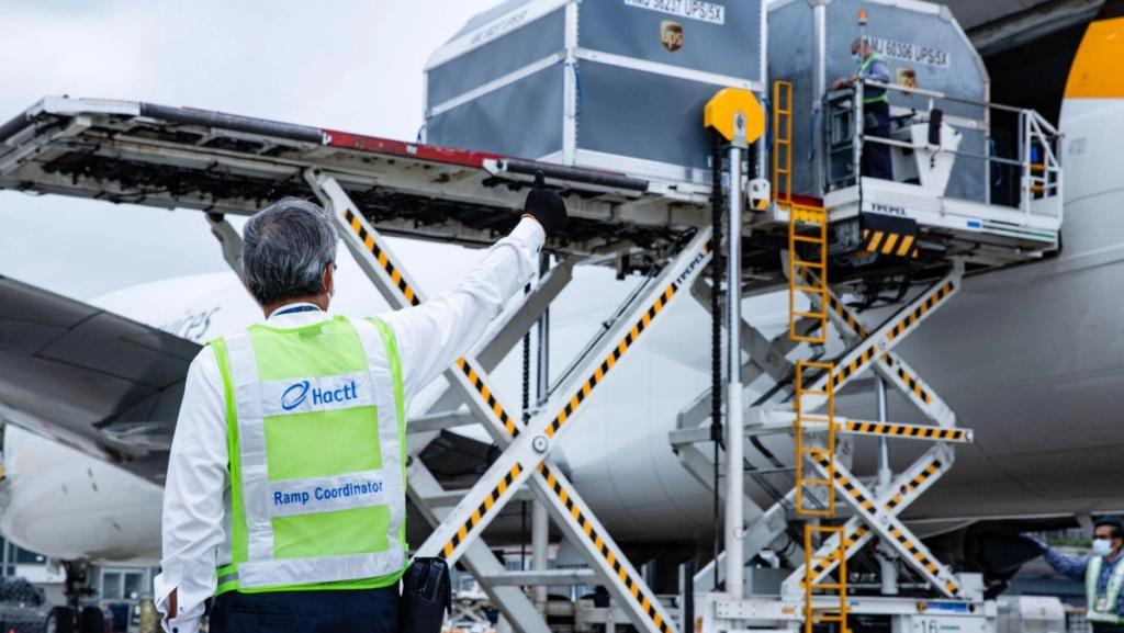 Hactl ramp coordinator in a yellow safety vest signals to a high-loader lifting a UPS container into a cargo aircraft on the tarmac