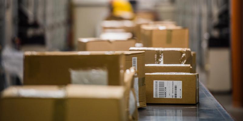 Cardboard parcels with shipping labels lined up on a metal conveyor belt in a warehouse, worker blurred behind.